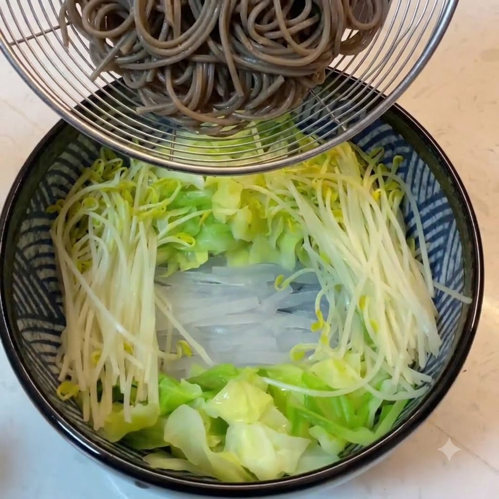 Cooked buckwheat noodles held in a wire strainer above the vegetable-filled bowl.