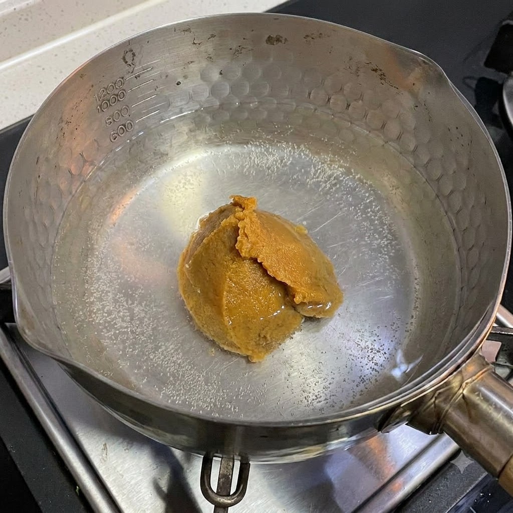 A spoonful of white miso paste dissolving in a pot of hot water on a stovetop.