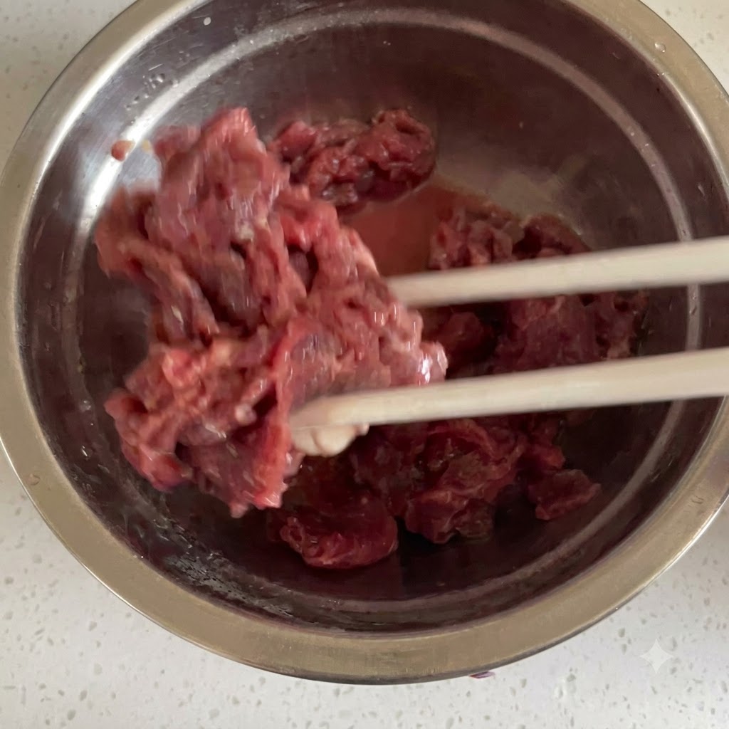 Thinly sliced raw beef being thoroughly mixed with marinade ingredients using chopsticks inside a stainless steel bowl.