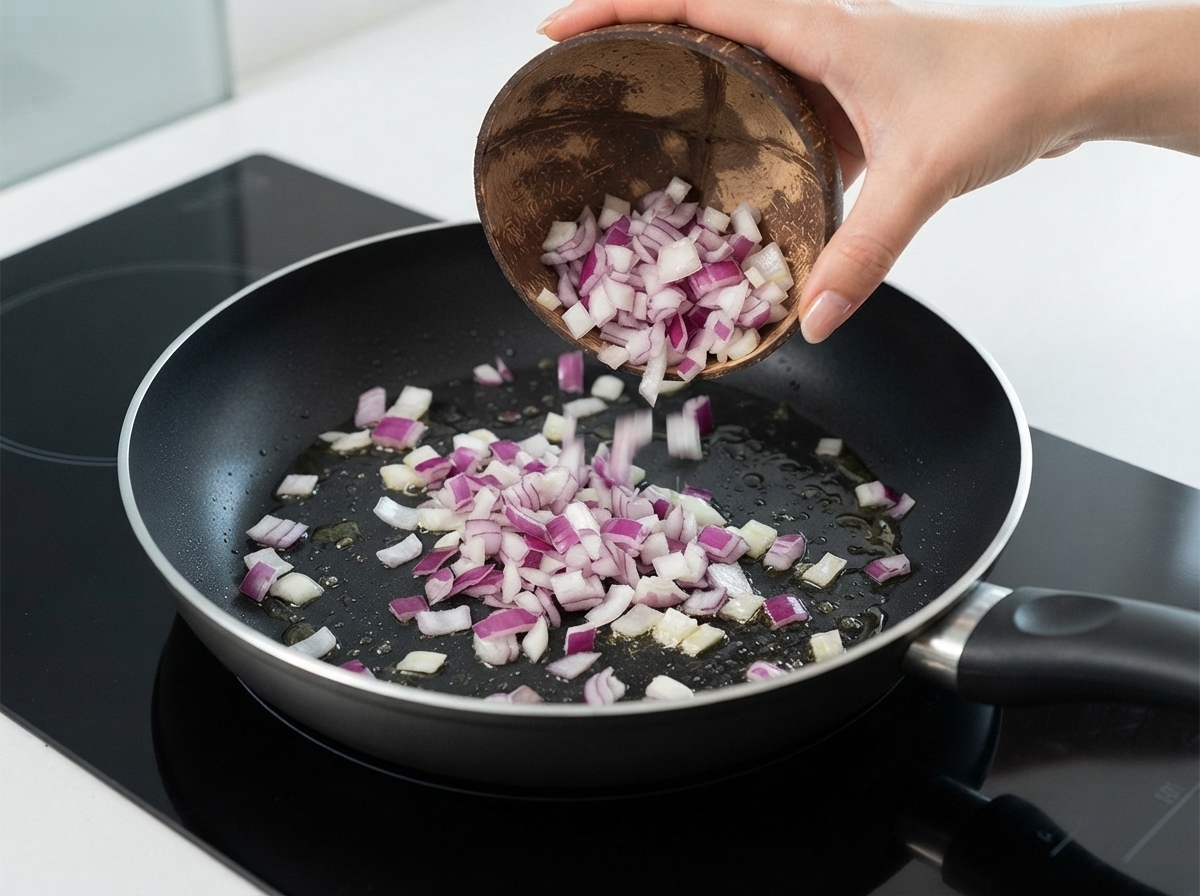Diced red onions being poured from a coconut bowl into a frying pan with oil.