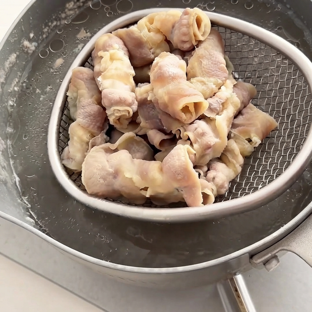 A wire strainer lifting parboiled beef rolls out from a pot of hot water.