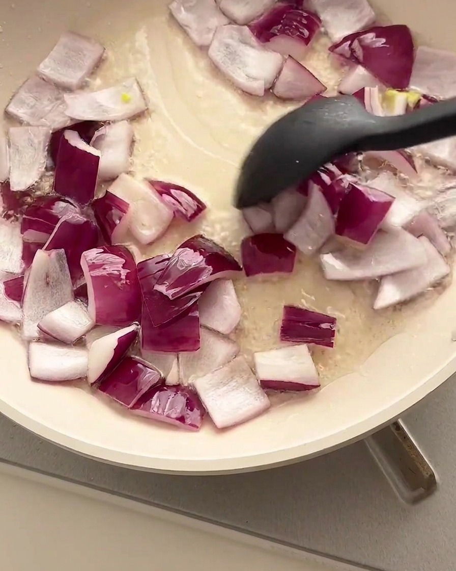 Diced red onions being stir-fried in an oiled pan using a black spatula.