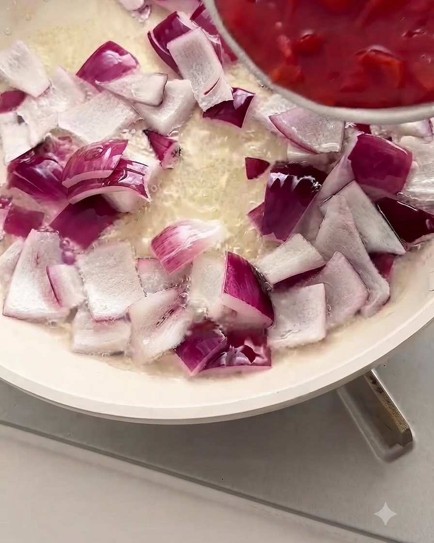 Chopped red tomatoes being poured from a bowl into a white pan filled with diced red onions and oil.
