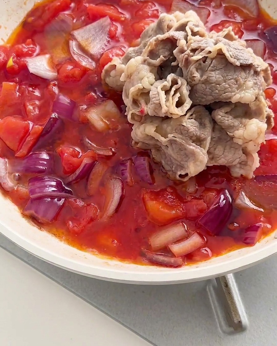 A pile of blanched beef rolls added into a pan with a simmering mixture of red tomatoes and onions.