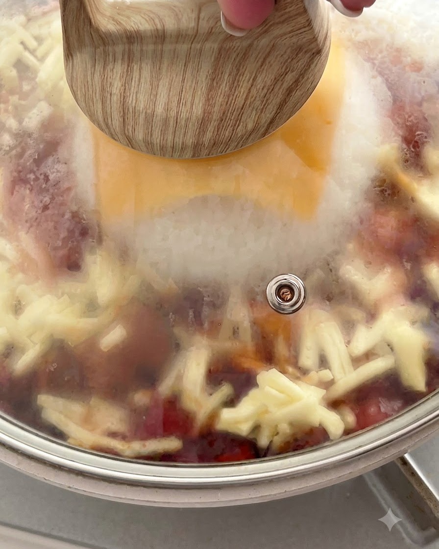 A pan covered with a glass lid showing steam trapped inside above the melting cheese and rice.