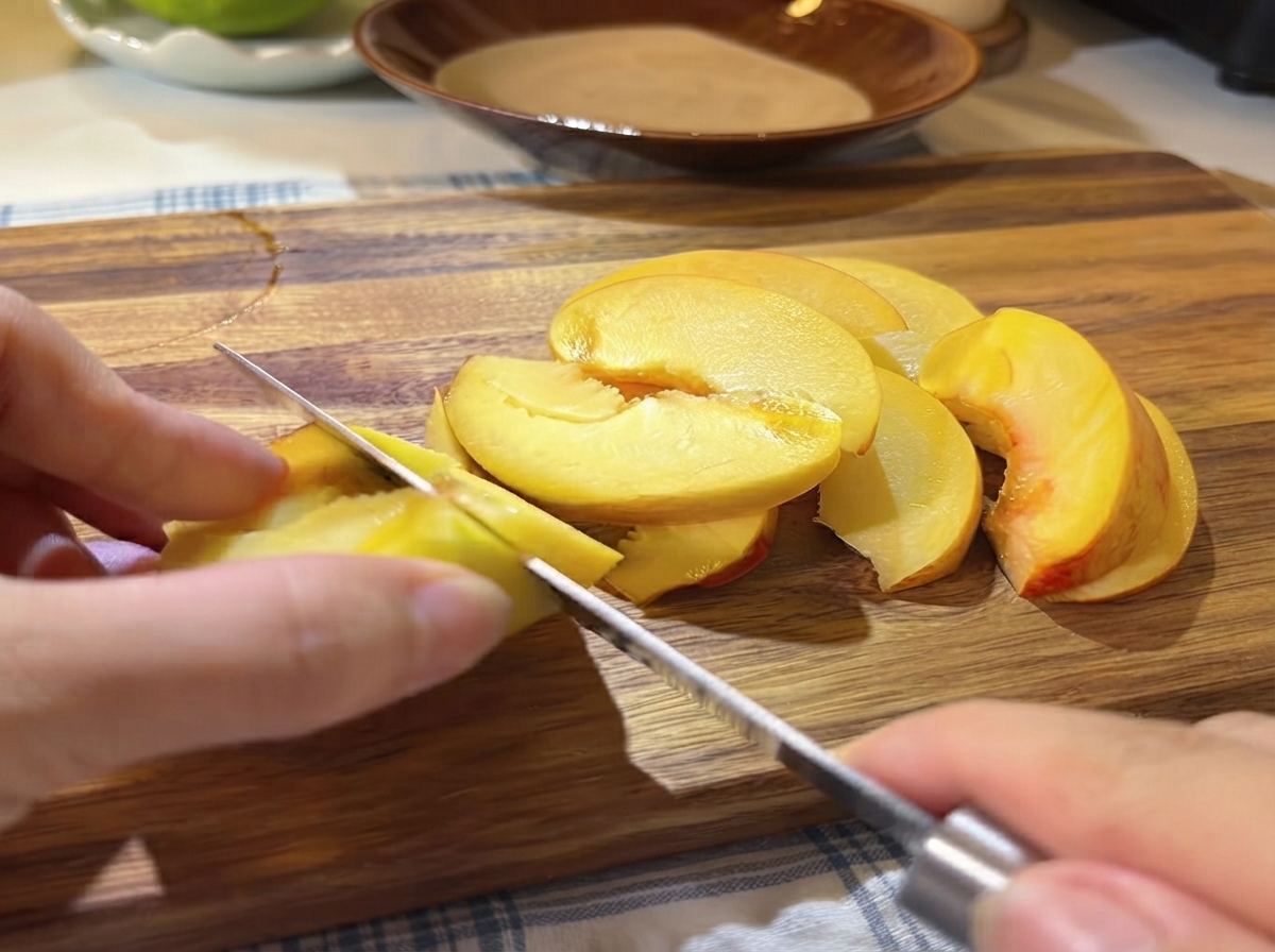 A hand using a small knife to slice fresh yellow peach halves into thin wedges on a wooden cutting board.