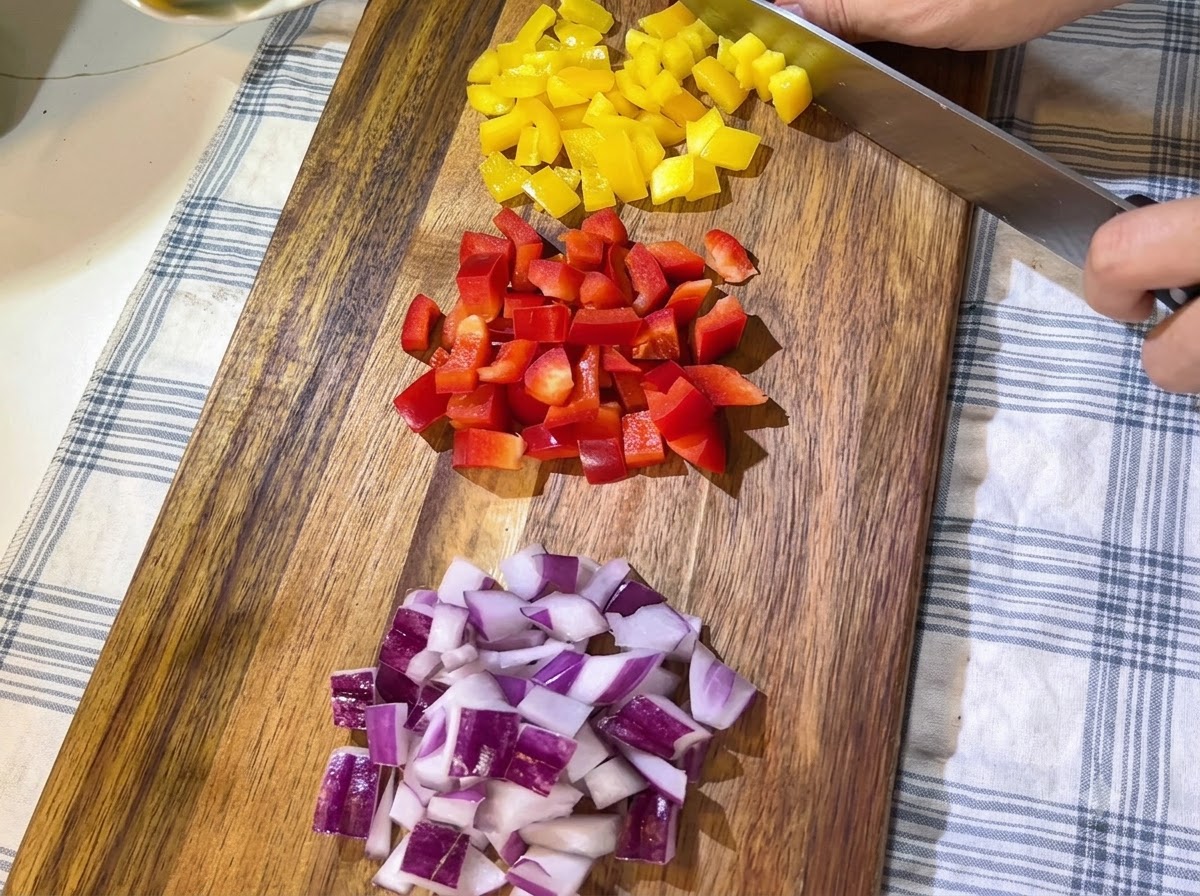 Piles of freshly chopped red and yellow bell peppers and diced red onions neatly arranged on a wooden cutting board.