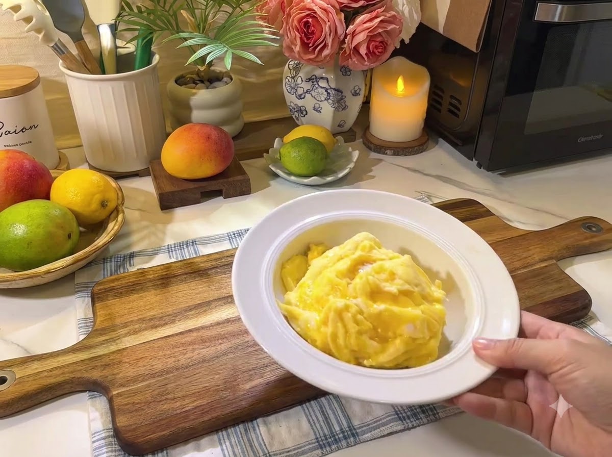 A plate of soft scrambled eggs resting on a wooden board next to fresh fruits and kitchenware.