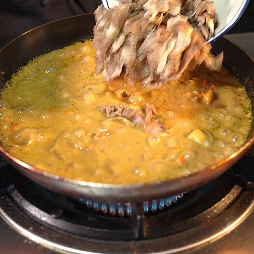 Pre-processed beef slices being poured from a white bowl into a simmering, thickened yellow curry sauce in a dark pan.