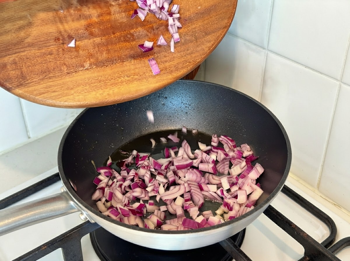 Chopped red onions being poured from a wooden board into a hot frying pan with oil.