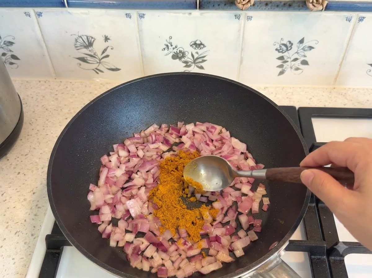 Curry powder being measured with a spoon and added to sautéed onions in a pan.