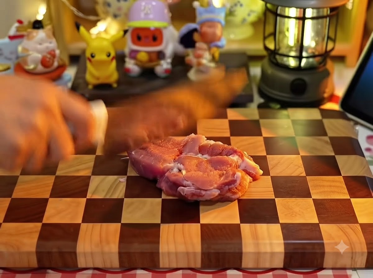 A hand using a kitchen knife to score a raw piece of pork meat on a checkered wooden cutting board.