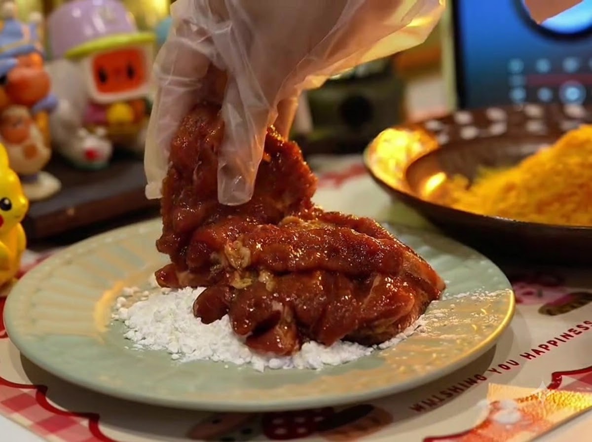 A gloved hand holding a piece of marinated pork chop over a bed of white starch on a pale green plate.
