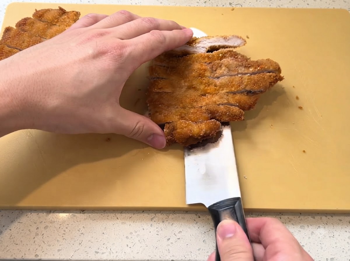 A golden-brown fried pork cutlet being sliced into strips with a sharp knife on a yellow cutting board.