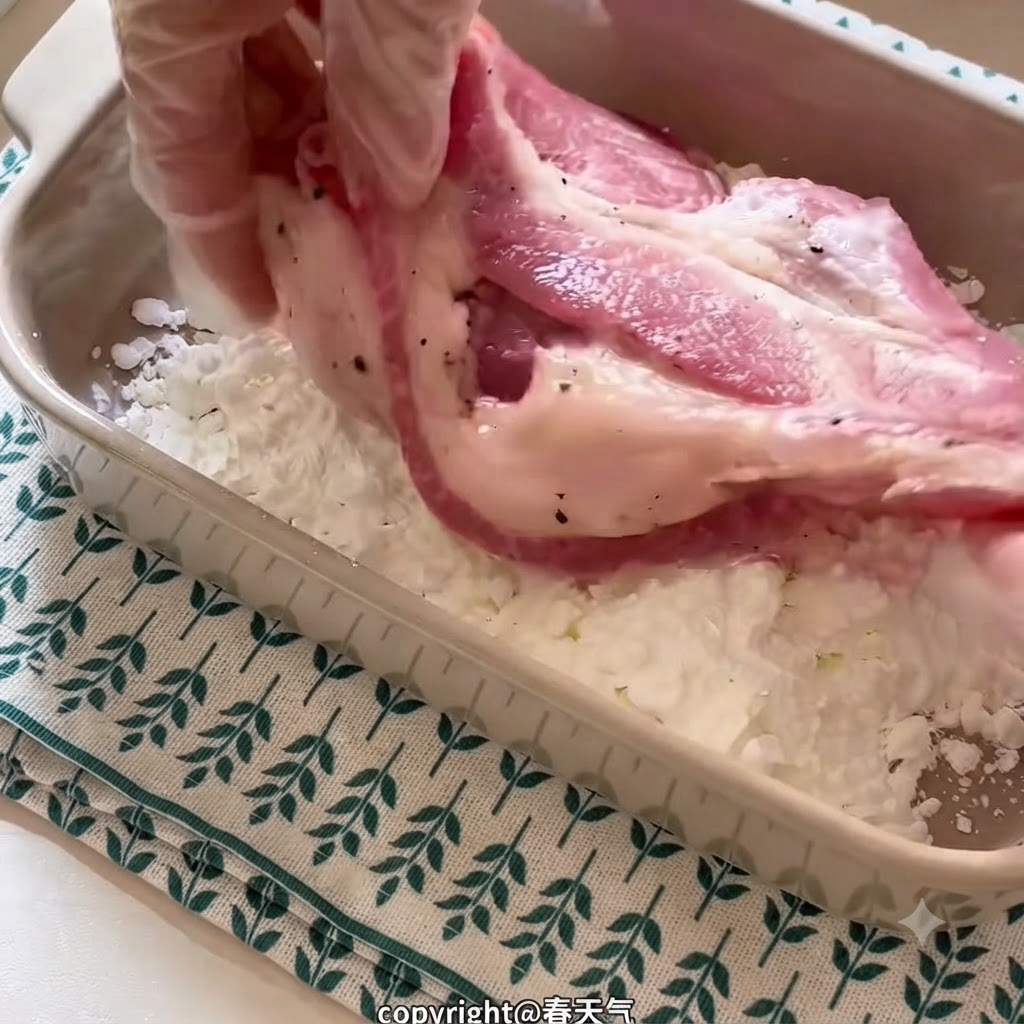 A gloved hand holding a marinated pork chop and coating it in starch inside a rectangular ceramic dish.