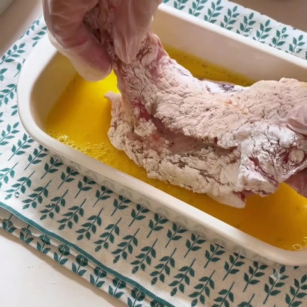 A gloved hand lifting a starch-coated pork chop out of a container of yellow beaten egg liquid.