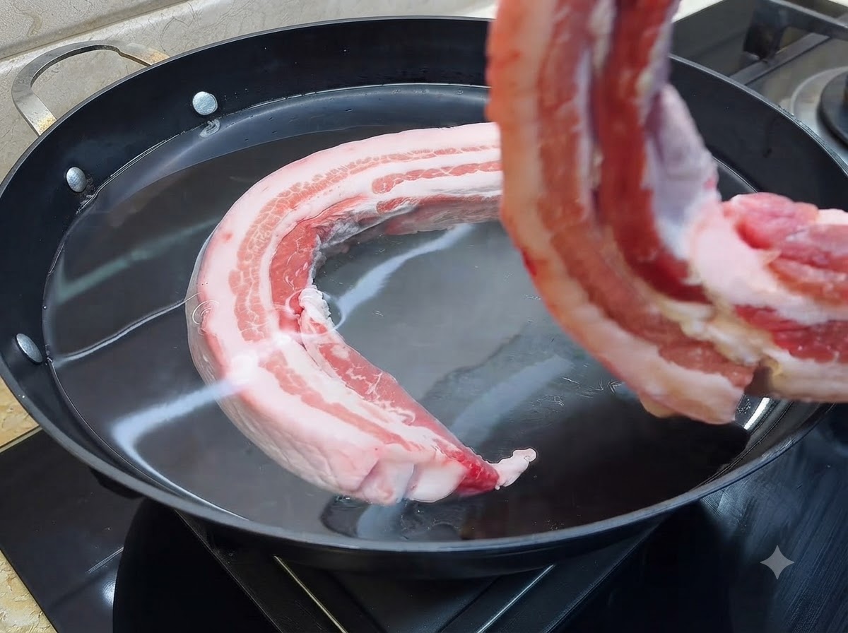 A piece of raw pork belly being lowered into a large pan filled with cold water.