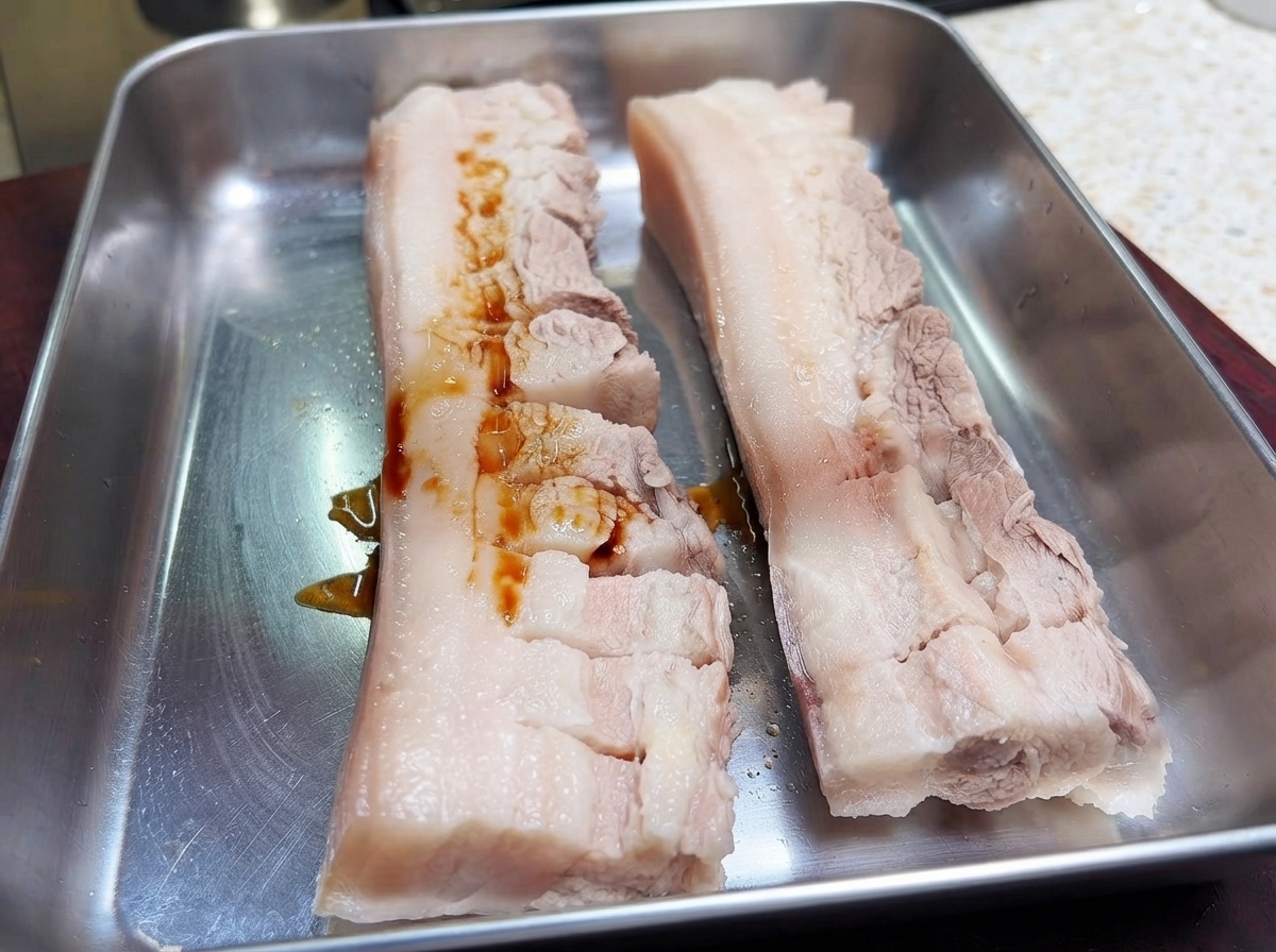 Two pieces of scored pork belly on a metal tray with dark soy sauce poured over the meat.