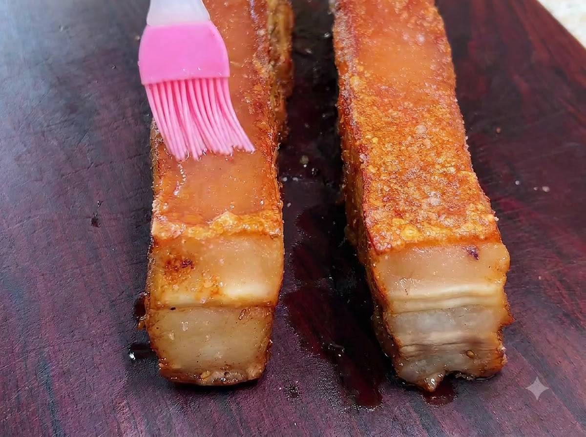 A pink silicone brush spreading white vinegar over the top surface of a roasted pork belly slab.