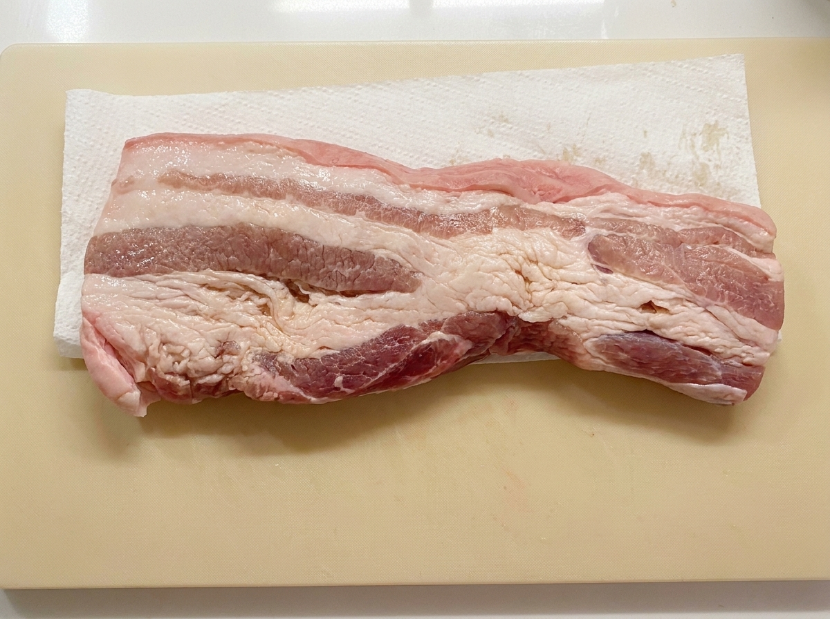 A pork belly strip lying flat on a paper towel on a cutting board while being dried.