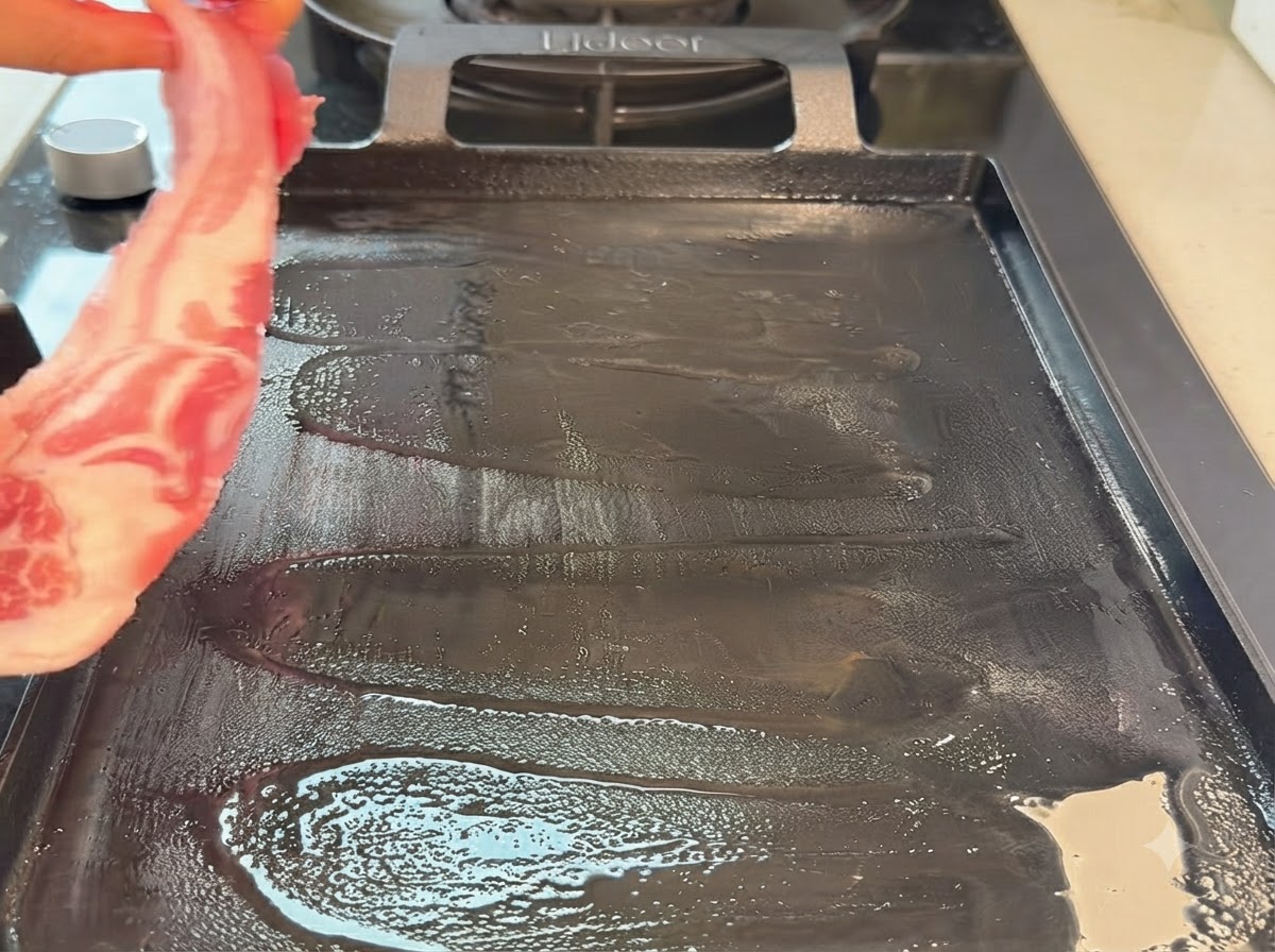 A raw pork belly strip being placed onto a hot teppanyaki grill pan.