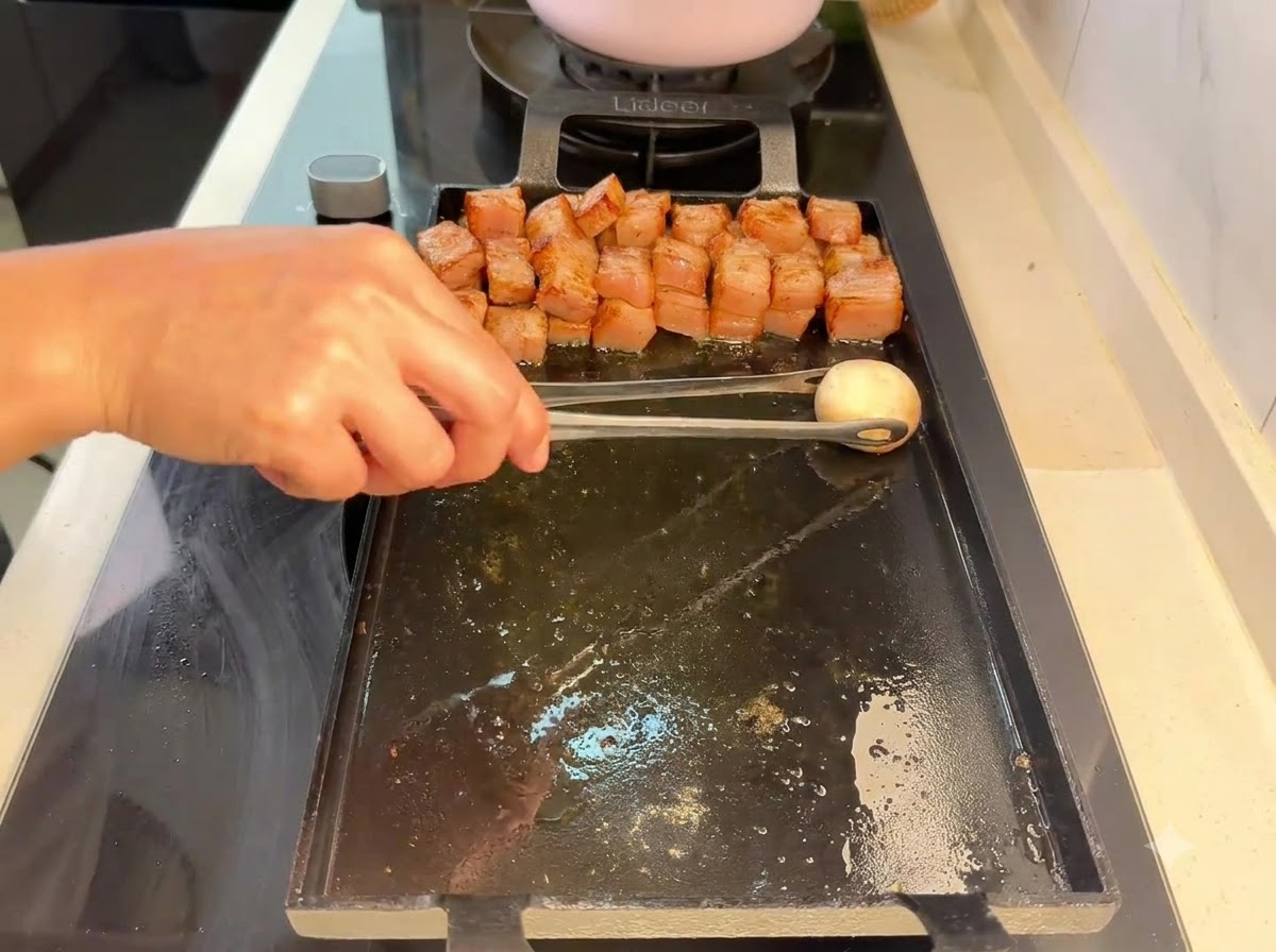 A button mushroom being placed with tongs onto the grill next to grilled pork pieces.