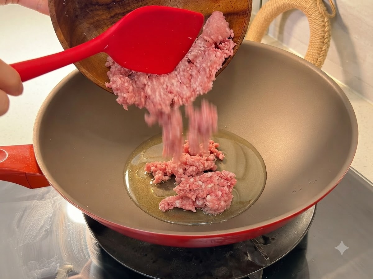 Fresh minced pork being poured from a wooden bowl into a heated red wok with oil.