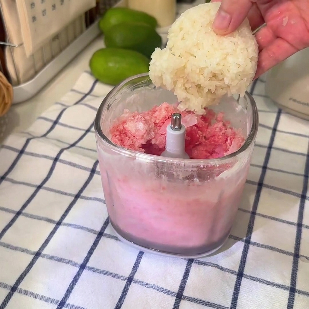 A ball of glutinous rice being dropped into a clear blender cup filled with pink watermelon slushie mixture.