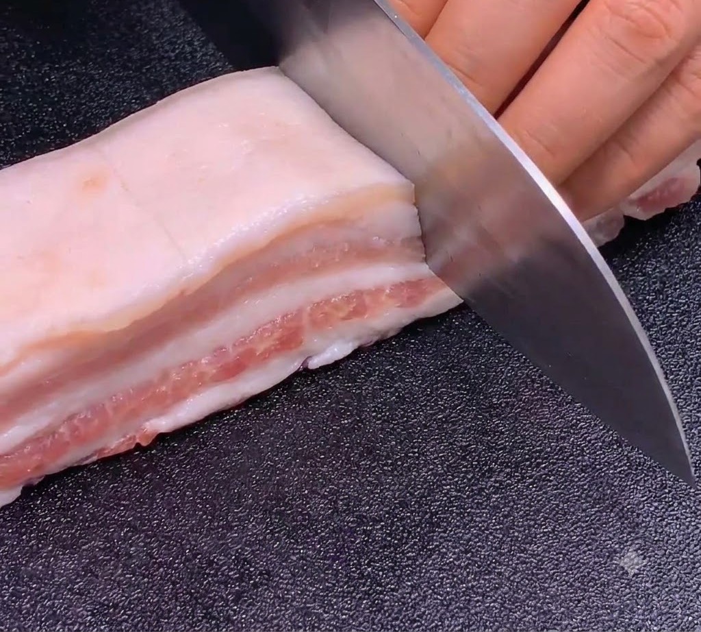 A sharp knife making precise, even cuts into a strip of pork belly on a dark cutting board.