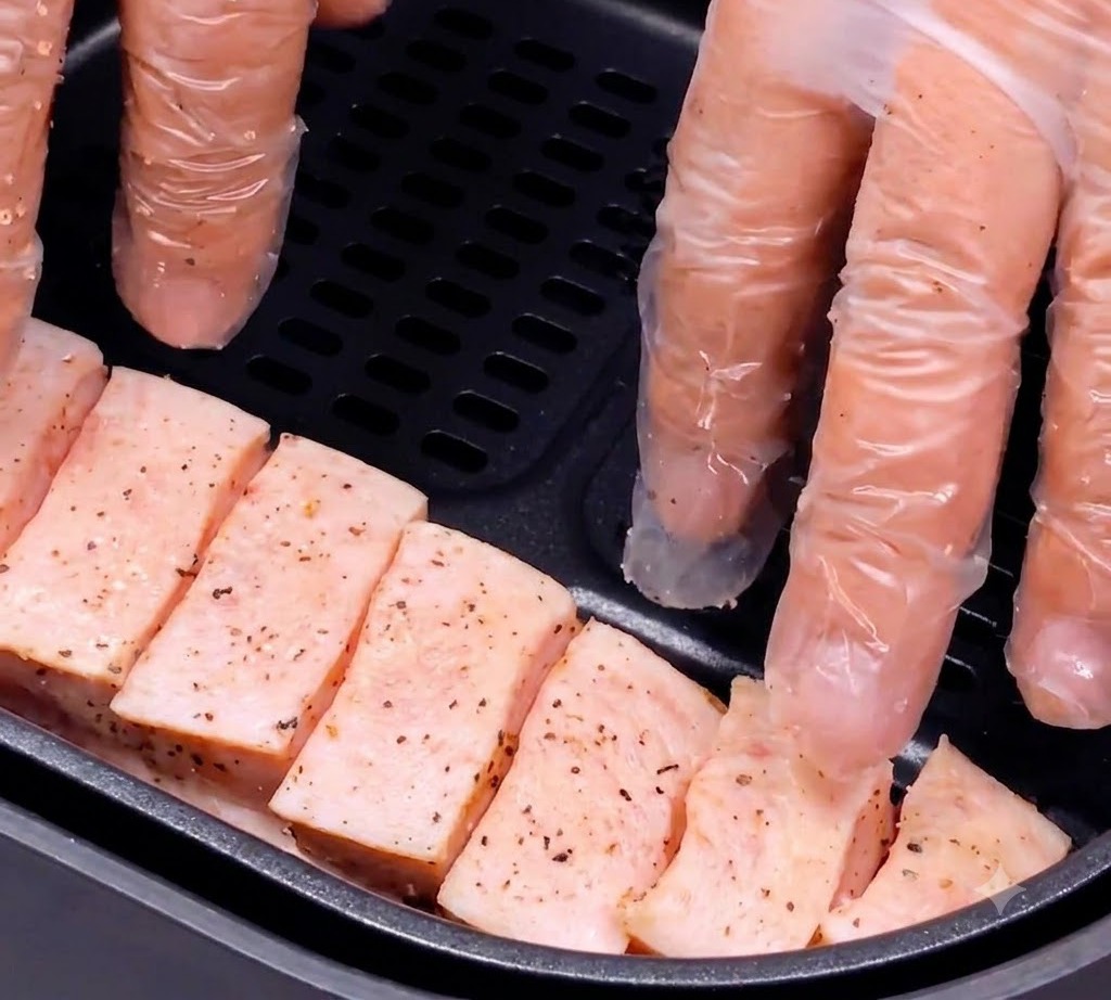 Seasoned pork belly strips being carefully placed into the dark basket of an air fryer by gloved hands.