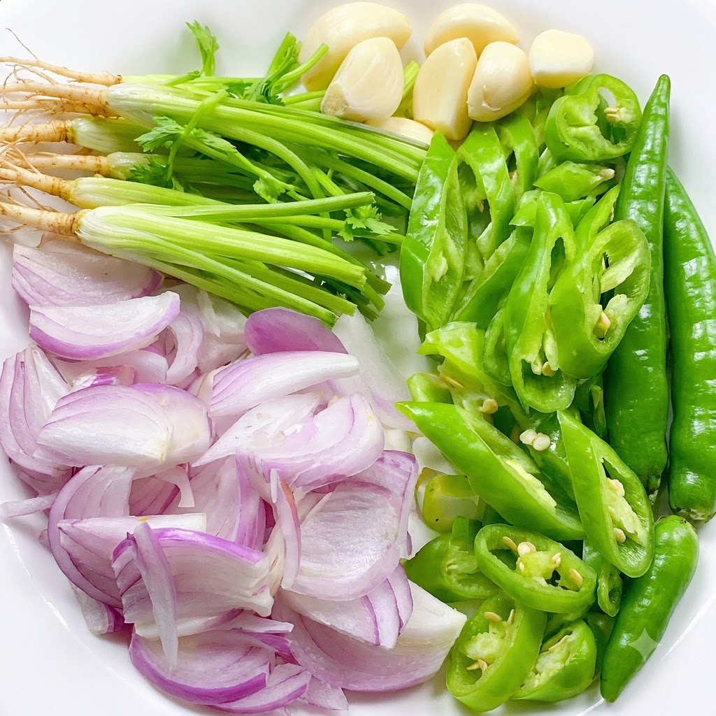 Fresh cilantro roots, sliced green chilies, sliced shallots, and whole garlic cloves arranged neatly on a white plate.