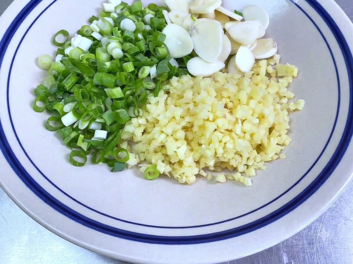 A white bowl containing finely chopped green scallions, sliced garlic cloves, and minced ginger.