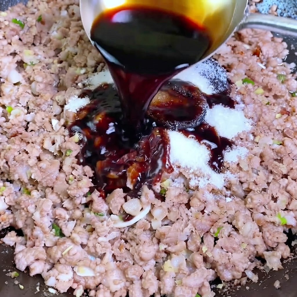 Dark soy sauce being poured from a silver ladle into a wok containing minced pork and white seasonings.