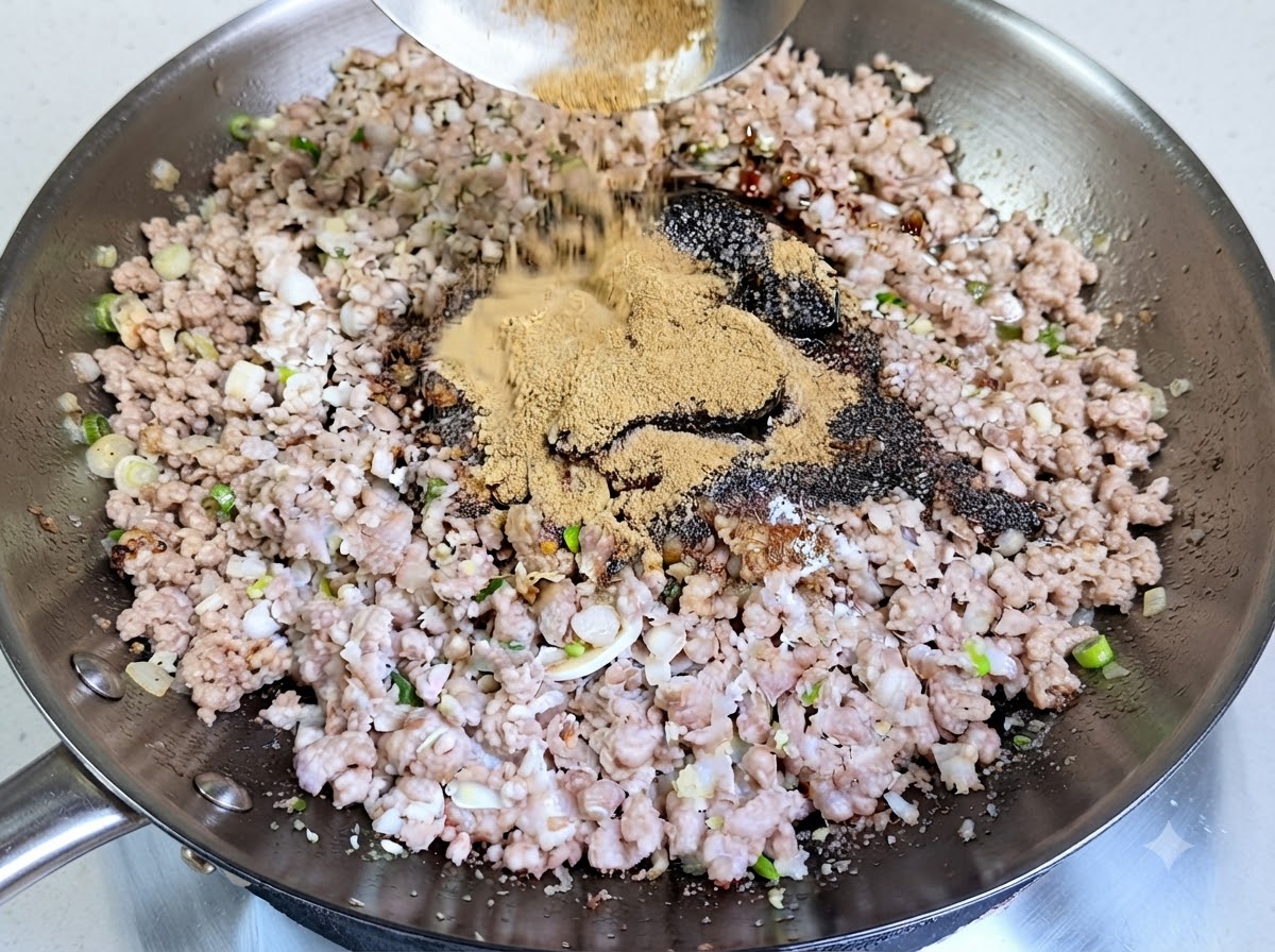 A generous heap of light-brown spice powder being poured from a ladle into the wok over seasoned minced pork.
