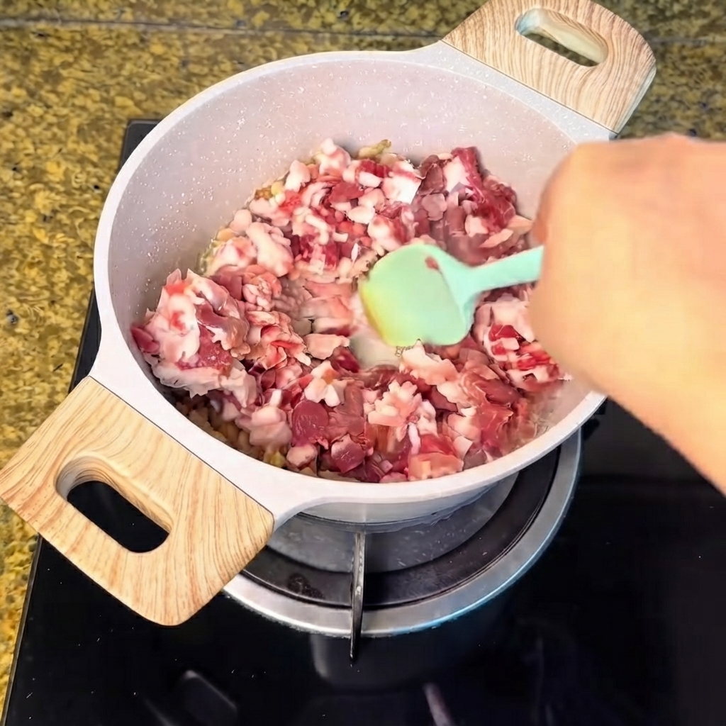 Diced pork meat being poured into the pot of rendered fat and mixed with a green spatula.
