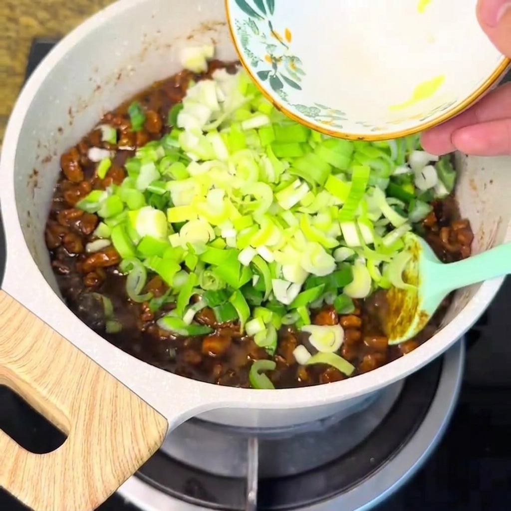 Chopped green onions being poured from a bowl into the bubbling meat sauce.