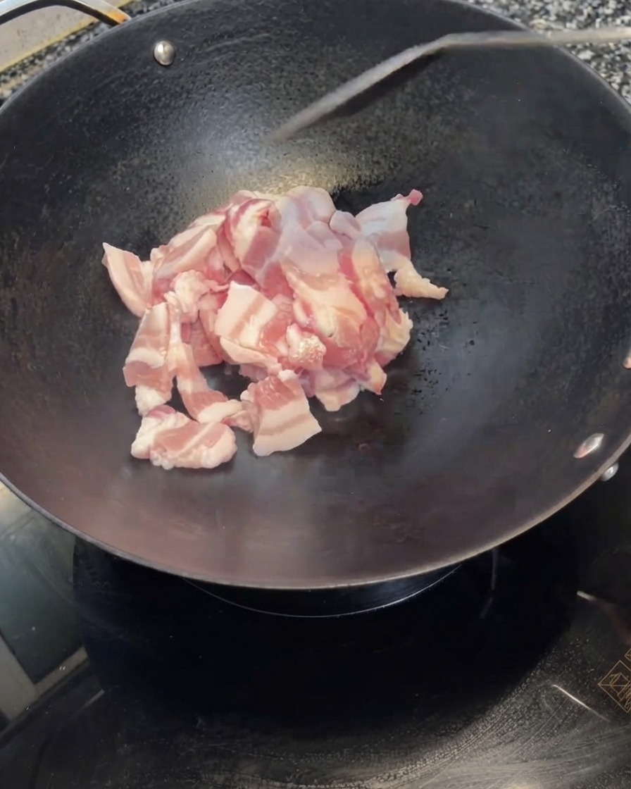 Sliced pork belly being tipped from a white bowl into a hot black wok.