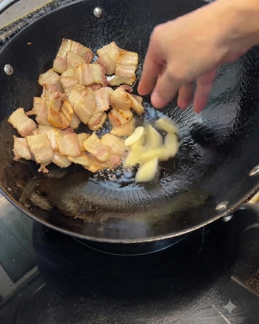 Smashed garlic cloves being tossed into a wok containing browned pork belly pieces.
