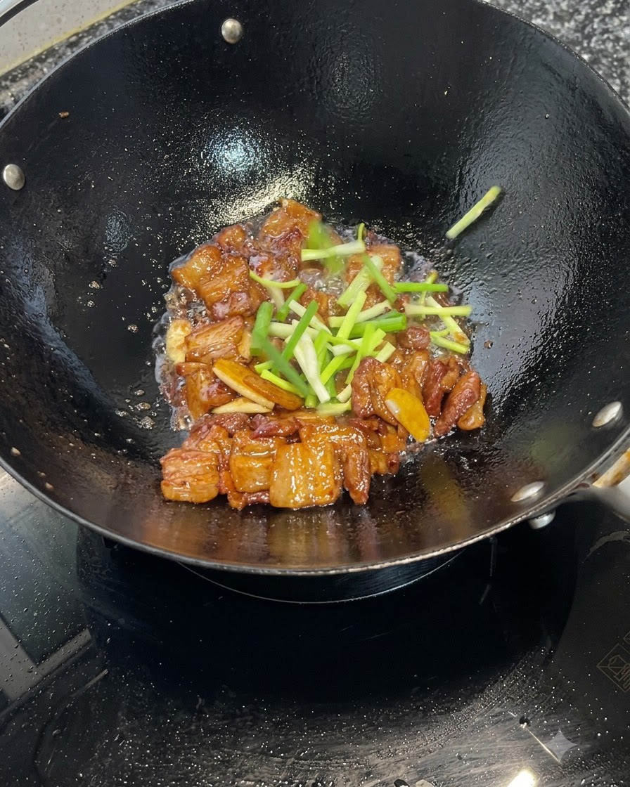 Fresh green onion sections scattered over caramelized pork belly pieces cooking in a wok.