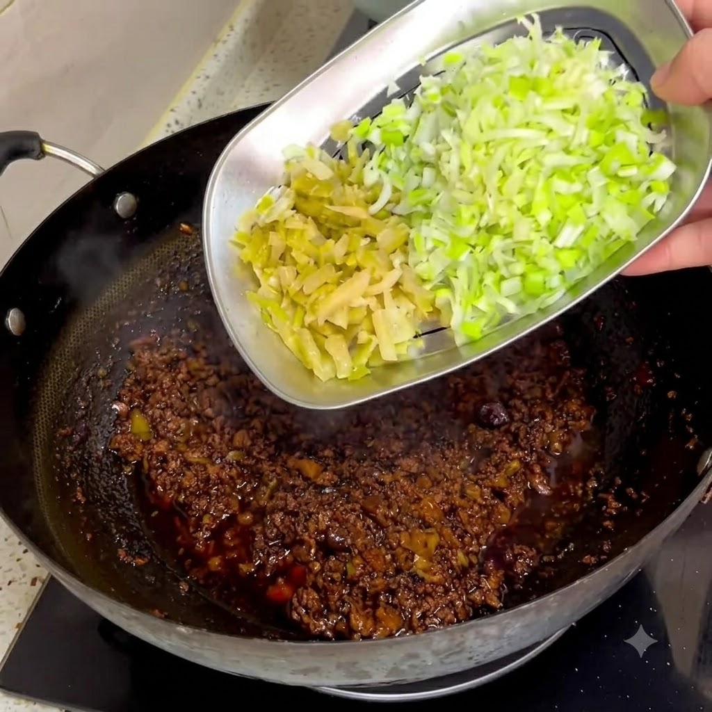Chopped preserved mustard greens and green onions being slid from a metal tray into a dark meat sauce in a wok.