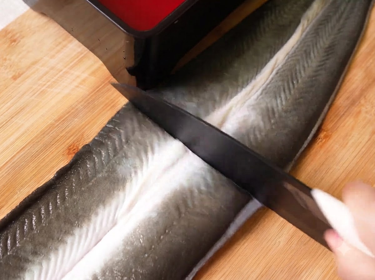 A sharp knife slicing a large butterflied raw eel into three sections on a wooden cutting board.