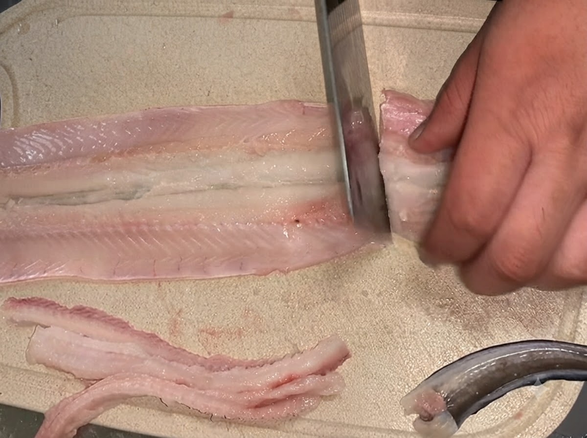 A knife cutting a deboned raw eel fillet into two separate sections on a cutting board.
