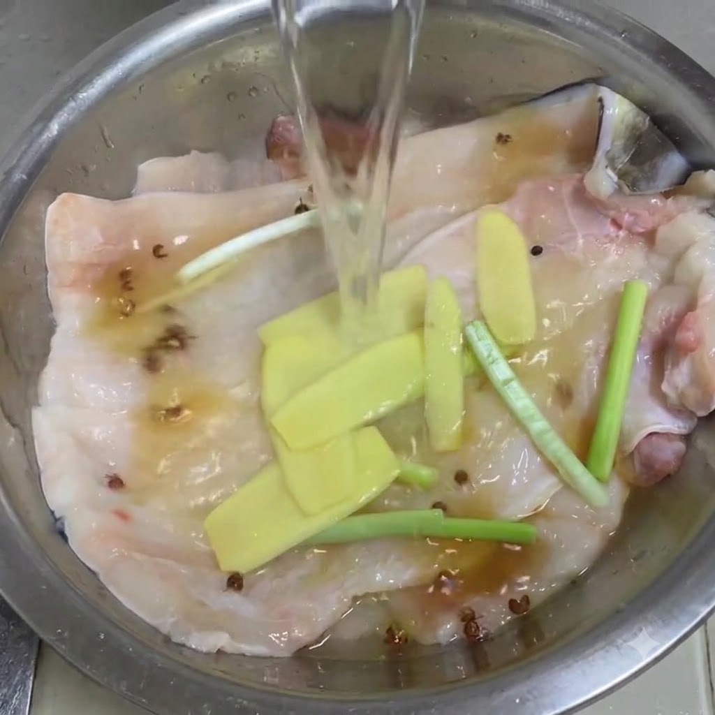 Clear water being poured into a metal bowl containing eel fillets, ginger, scallions, and Sichuan peppercorns.