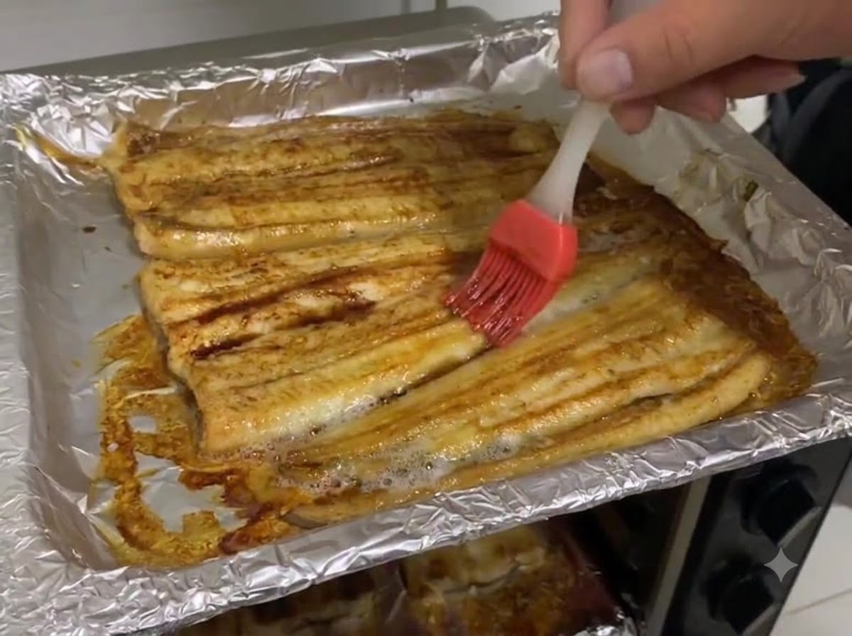 Dark soy sauce being brushed onto the surface of baked eel fillets in a foil-lined baking sheet.