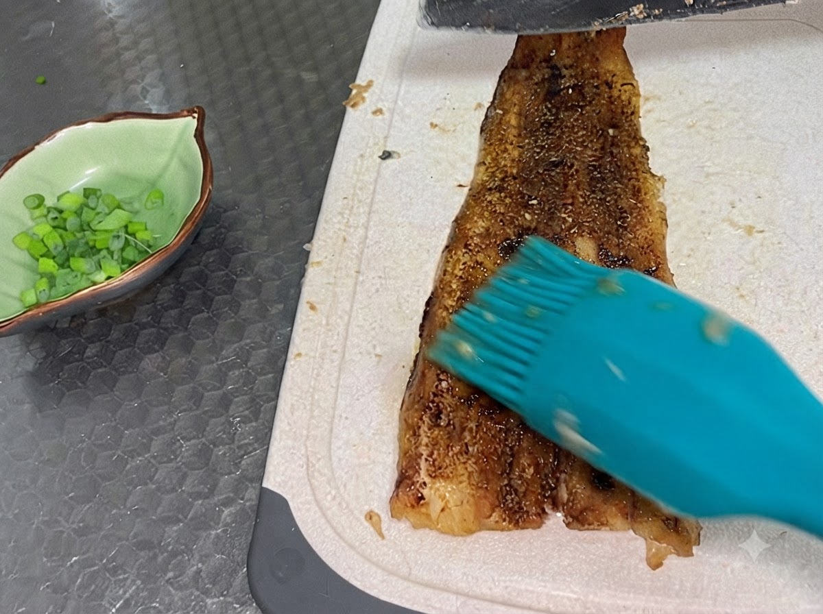 A fully baked eel fillet being sliced on a white cutting board next to a small green bowl filled with chopped scallions.