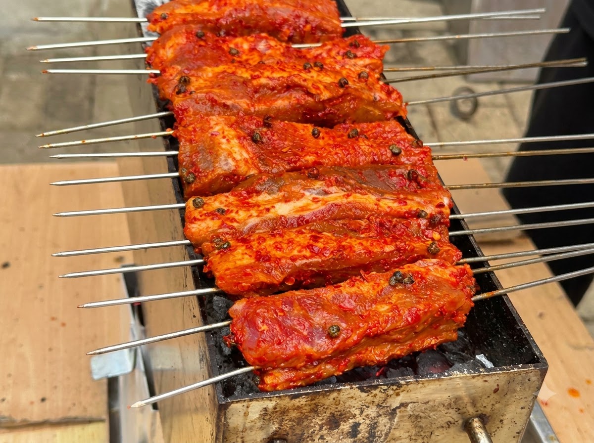 Multiple skewers of red-coated ribs lined up and grilling on top of a charcoal pit.