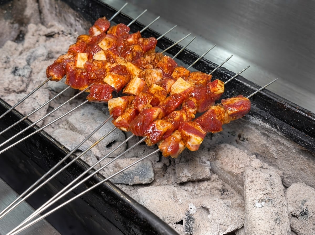 Multiple metal skewers of seasoned pork meat arranged neatly over a burning charcoal grill.