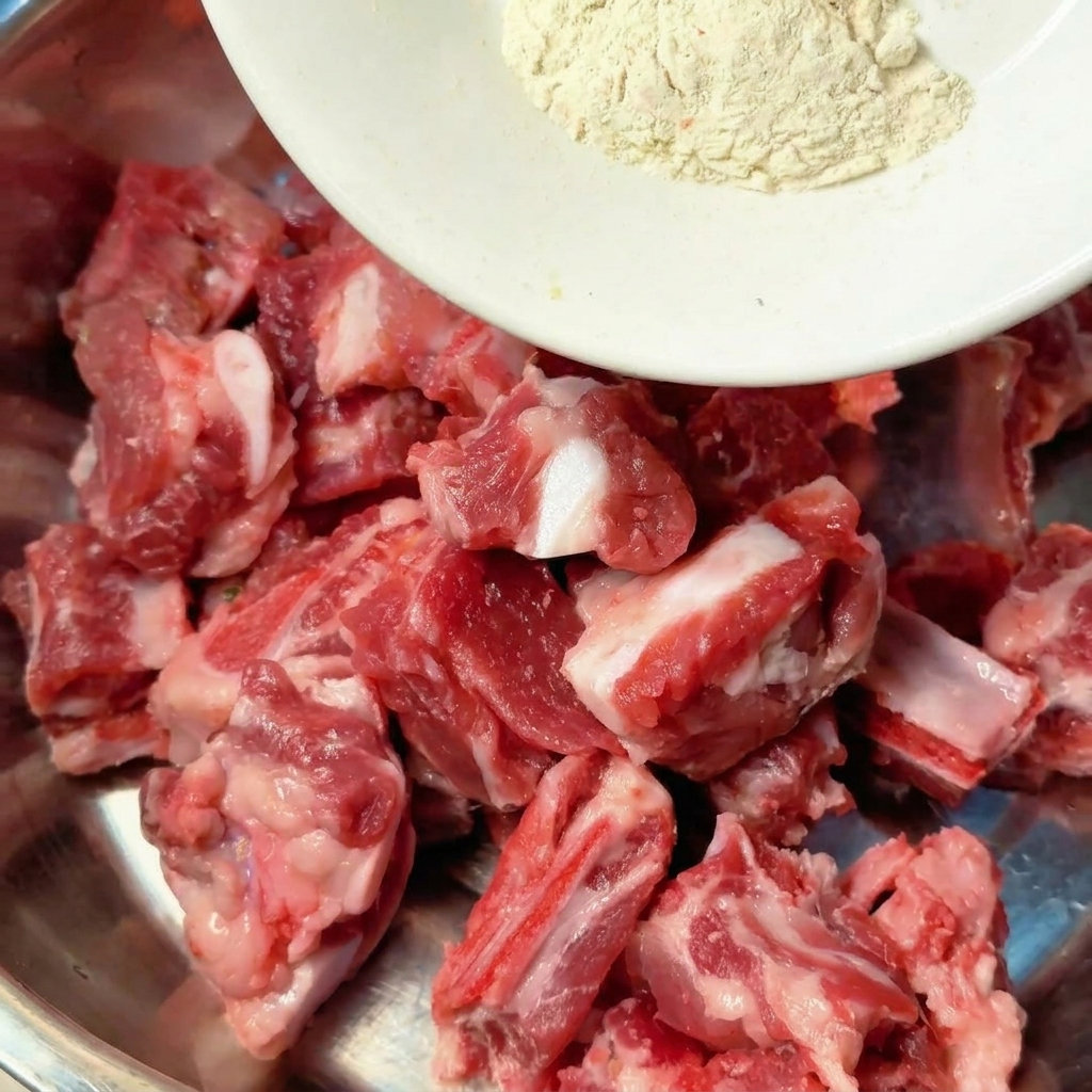 Spices and liquid seasonings being poured directly onto raw pork ribs in a metal mixing bowl.