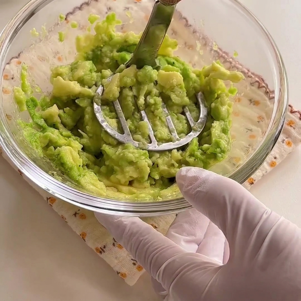 Avocado halves being mashed into a vibrant green paste inside a glass bowl with a metal masher.