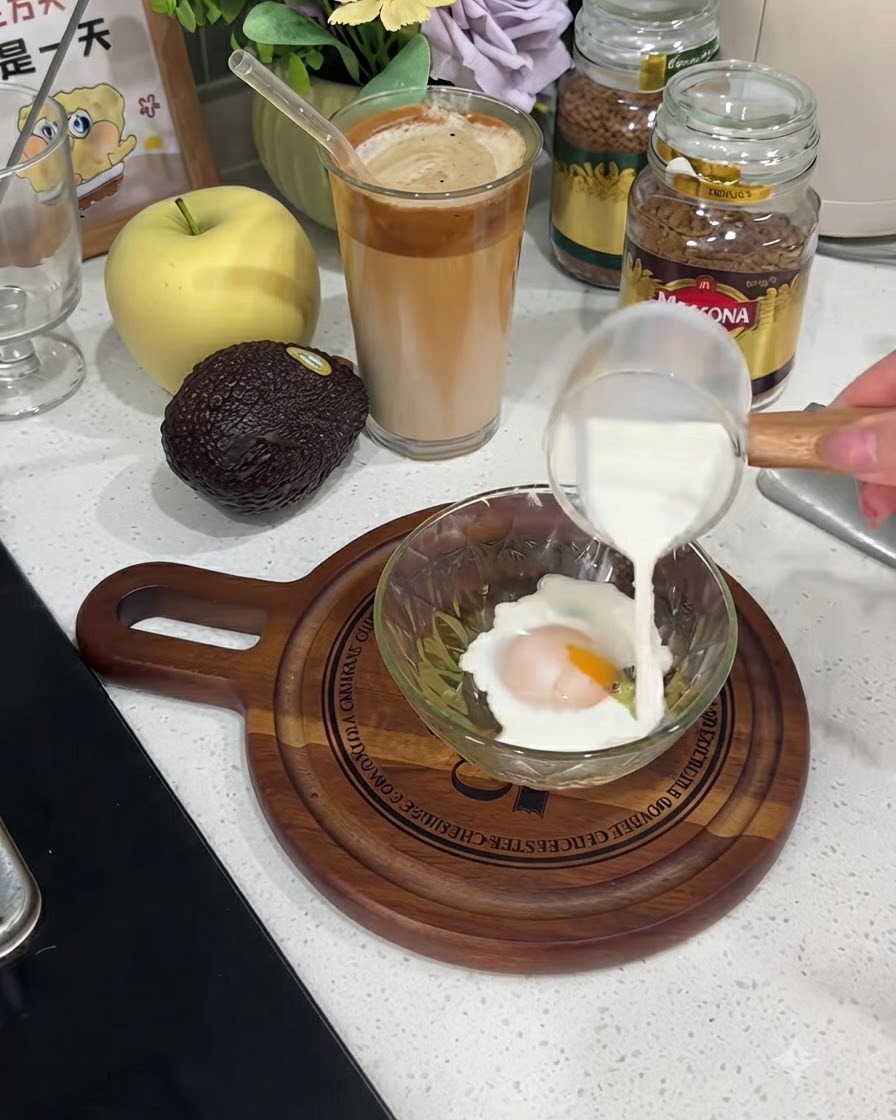 Milk being poured from a small glass pitcher into a bowl containing a cracked egg.